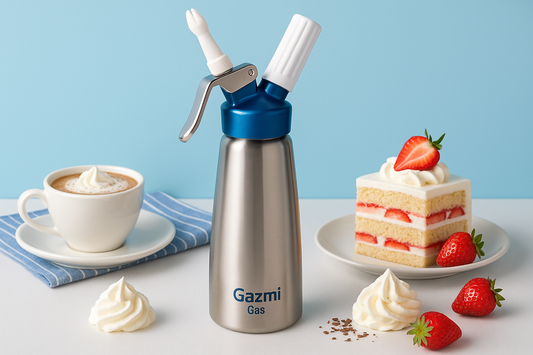 A sleek stainless steel whipped cream dispenser branded with Gazmi Gas, placed on a white table with a cup of cappuccino topped with whipped cream and a slice of strawberry shortcake against a soft blue background.