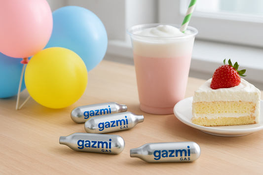 Gazmi Gas nitrous oxide canisters neatly arranged on a light wooden table, surrounded by colorful balloons, a sparkling drink, and a slice of strawberry cake. Servicegazmi